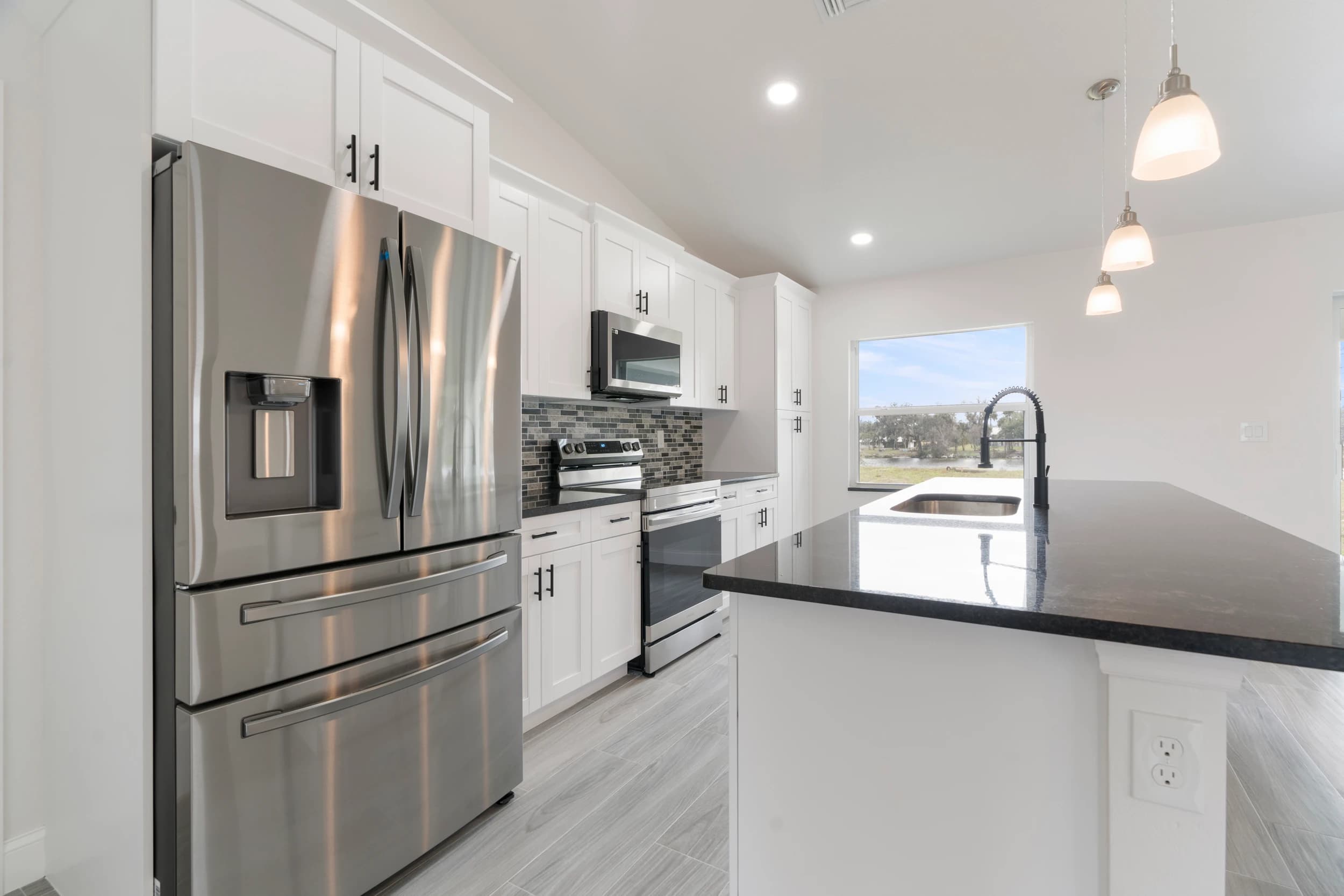 Stylish modern kitchen in a newly built custom home by Vreeland Roofing in Florida. Features include white shaker cabinets, stainless steel appliances, a sleek black quartz countertop island, and pendant lighting. Open-concept design with large windows allowing natural light, creating a bright and spacious cooking area.